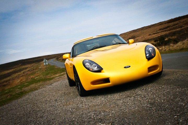 Nice Car Photo locations Middlesbrough Area? - Page 1 - North East - PistonHeads - This image features a **yellow sports car** parked on a **gravel lot** with a scenic backdrop. The car is an **F350**, characterized by its sleek design, specific to the **Ford Mustang** model. The gravel lot appears to be located near a **mountain range**, adding a dramatic element to the scene. In the background, there's a solitary figure who seems to be inspecting the car or taking photographs. The sky overhead is a clear **blue**, suggesting it might be a sunny day.