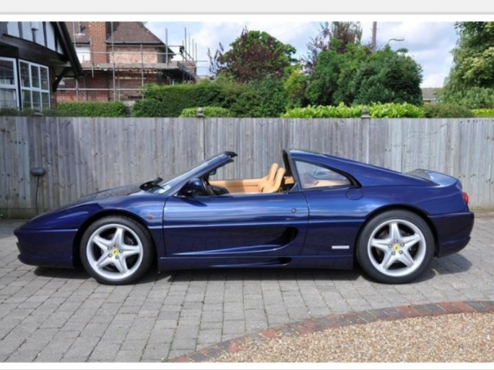 355 A future Classic (2014)  - Page 1 - Ferrari V8 - PistonHeads - The image shows a classic blue Ferrari sports car parked adjacent to a wooden fence. The car boasts a convertible top and features gold rims and a yellow center on the rear wheels. It's positioned under a partly cloudy sky, bathed in natural light that accentuates its glossy finish. A brick driveway adds to the setting, suggesting a private residence or a similar location.