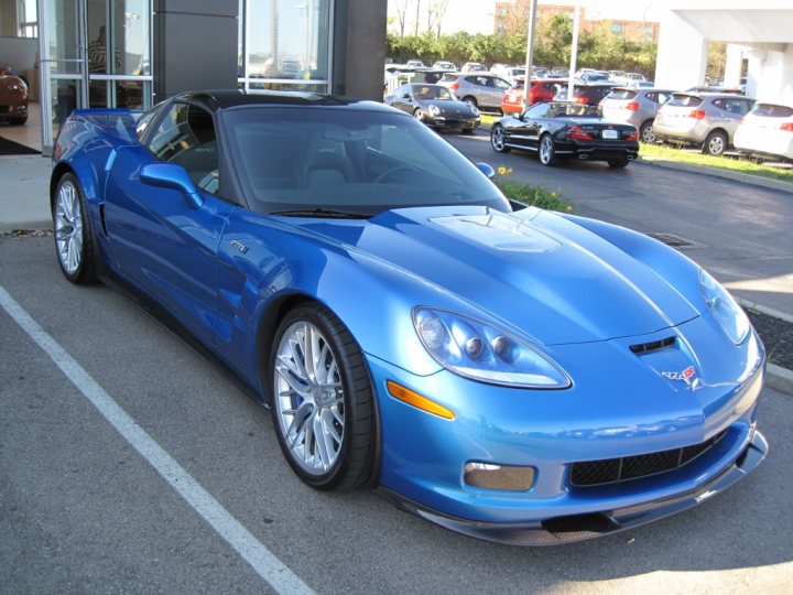 I finally gave in....... - Page 1 - Corvettes - PistonHeads - The image shows a vivid blue Corvette parked in a brightly lit area. The car is positioned in the foreground and is the central focus of the photo. It is during the day, and the lighting highlights the glossy finish and sleek design of the vehicle. The background consists of a softly blurred array of other parked cars, with one notably darker. The setting appears to be a dealership or car show, characterized by its open nature and ample outdoor space.