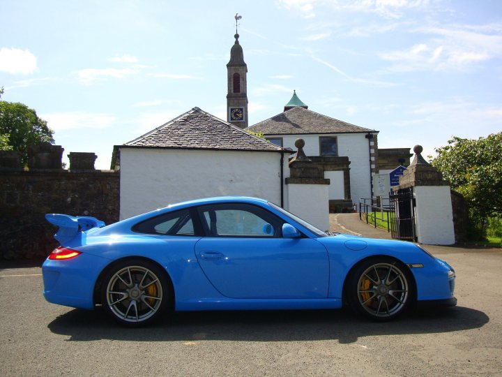 Show off your GT, past and present... - Page 12 - 911/Carrera GT - PistonHeads - The image depicts a vibrant blue Porsche sports car parked in front of a traditional white building with a sloping roof. This is a model Porsche with a rear spoiler and angular body design, typical of many performance-oriented automobiles. The sunlight casts shadows behind the car, indicating it might be late afternoon. There is a solitary figure in the background, adding a touch of life to the scene. The sky above is a clear blue, suggesting a sunny day. There are no visible texts or distinguishing marks that provide additional context to the location or event. The image conveys a sense of serenity and elegance, with the car being the main focal point.