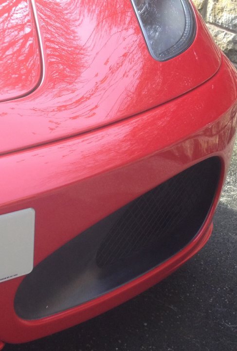 A red and black motorcycle is parked in a parking lot - Pistonheads - The image features a close-up of the front of a bright red car, captured from an angle that emphasizes the round headlight positioned below a smaller air intake. The reflective surface of the car hood displays subtle lines and reflections indicative of its polished and glossy finish. The soft focus and limited background detail bring the viewer's attention to the textured fingerprint-like surface of the red paint. The vehicle's unique curvature and design suggest it is a sports car.