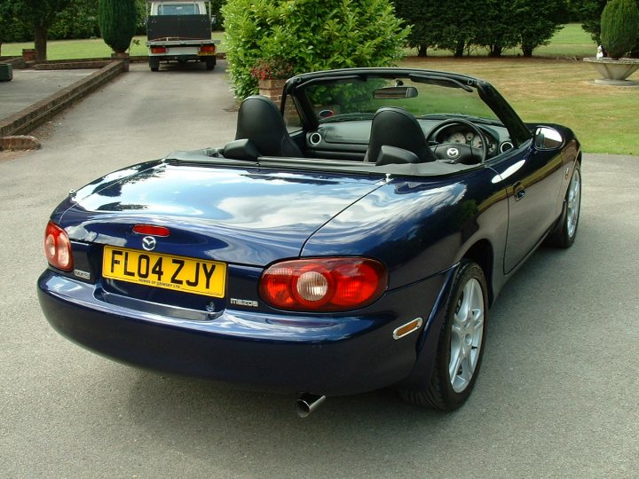 Pistonheads - The image showcases a dark blue two-door convertible parked on the side of the road. The car is equipped with a soft top and features a small red taillight. The vehicle registration indicates it belongs to a person named Jobe FLO4C, and the road curves to the left in the foreground, suggesting it leads through a residential area.