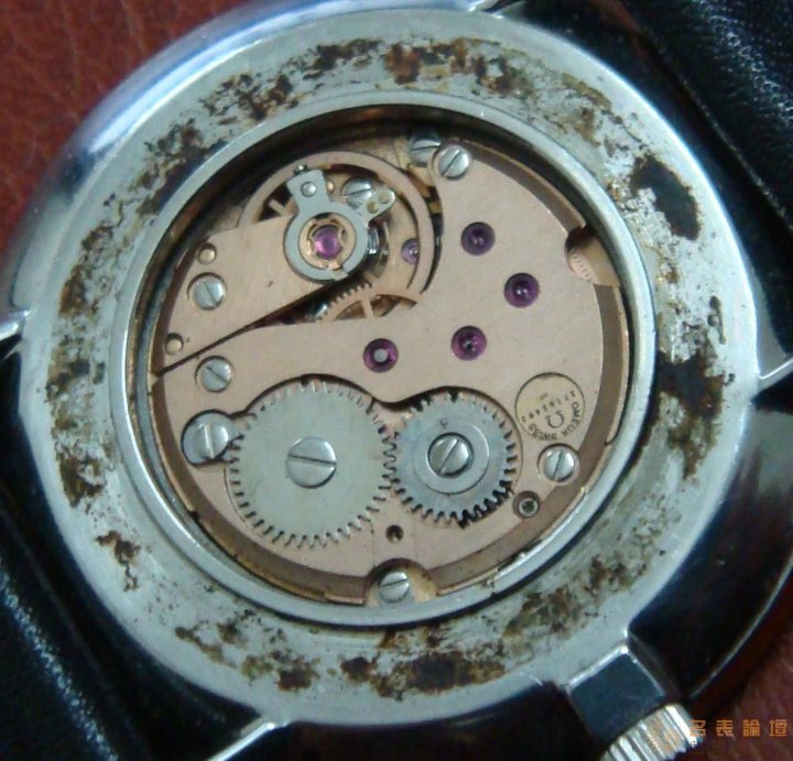 Model Pistonheads Omega Ville Case - The image features a close-up view of the inner workings of a mechanical watch. The intricate mechanism includes several gears and components, predominantly made of metal with hints of pink, indicating possible synthetic materials or a decorative aspect. The overall condition appears to be somewhat worn, with rust or dirt accumulation visible on various parts of the watch. This gives the image a gritty, industrial feel that emphasizes the underbelly of this timepiece.