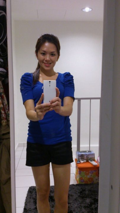 A woman holding a cell phone up to her ear - In the image, a young woman is the main focus, standing in a room within a mirror that reflects an interior view. She is dressed in a short-sleeved blue top and black shorts, holding a smartphone in her hands. A smile graces her face, suggesting she is in a pleasant mood. The room around her appears to be a living area or bedroom, evidenced by the presence of clothes and books in the background.