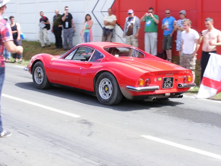 Pistonheads Classics - The image captures an exciting scene on a road where a vintage, red race car is the center of attention. A crowd of spectators, standing on both sides of the road, watches the car with interest. The car is driving on the right side of the road, indicating that the photo likely represents the European tradition of driving on the left side of the road. The spectators appear to be enjoying the event, suggesting it is a public event or race. The road itself is clean and well-maintained, extending towards a white building visible in the background.