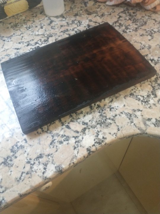 The bbq photo & recipe thread - Page 210 - Food, Drink & Restaurants - PistonHeads UK - The image shows a kitchen counter with a dark, possibly maple wood cutting board placed on it. The countertop appears to be made of marble with white and grey veining. A bottle of what could be oil or cleaner is visible next to the cutting board. There's also a cabinet door beneath the countertop that seems to have been recently painted or refinished in white, contrasting with the dark wood cutting board above it. The overall scene suggests someone has prepared for cooking or meal preparation.