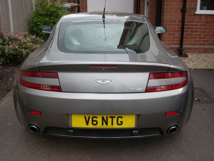 Pistonheads - The image shows a silver Aston Martin DB7 from the back. The car is parked on a short driveway. The registration plate of the car is clearly visible and reads V6 NTG. The body of the car reflects some sunlight, highlighting its well-maintained appearance. The tail light is red with a clear glass backing, allowing for visibility.