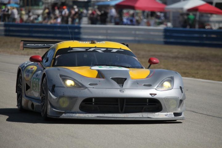 New SRT Advert - Page 1 - Vipers - PistonHeads - The image captures a thrilling moment on a race track, with a sports car in the foreground. The car is a vibrant mix of yellow and grey with a distinct red accent on the top edge of the front. It's a performance vehicle, specifically designed for racing, judging by its aerodynamic body work and large, bright lights on the front. In the background, there are multiple spectators or other participants lined up in front of tents, adding to the atmosphere of a racing event. The car is in motion, driving around a bend, which gives the impression of speed and competitive racing.