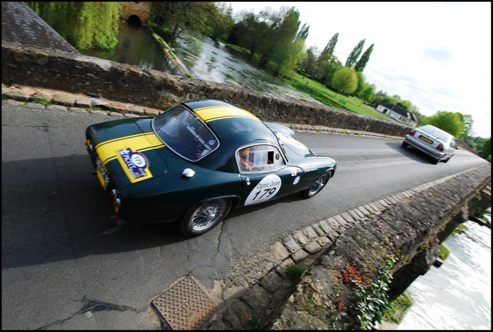 The Tour Auto came to town - Page 1 - Classic Cars and Yesterday's Heroes - PistonHeads - The image showcases a vintage green car adorned with yellow and white stripes, decorated with an identification number, 177. It's positioned on a narrow street, adjacent to a sturdy stone bridge that spans over a gentle, flowing river. The car is captured mid-journey, being driven past a house nestled within the verdant surroundings. In the background, you can observe more of the picturesque landscape, including a lush green lawn and trees, painting a serene and idyllic scene of a tranquil village.