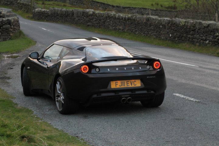 black evora spotted - Page 1 - Evora - PistonHeads - The image depicts a black sports car in motion on a road. The vehicle appears to be a Lotus, as indicated by the text on its license plate, which reads "FIJ EVC." The design of the Lotus is sleek and low to the ground, suggesting speed and agility. The car is traveling on a two-lane road, with a clear lane for the car visible in the image. The road is lined with a stone wall on its left side, indicating a boundary or property line. The background shows a rural landscape with rolling hills and trees, giving the setting a serene and peaceful atmosphere.