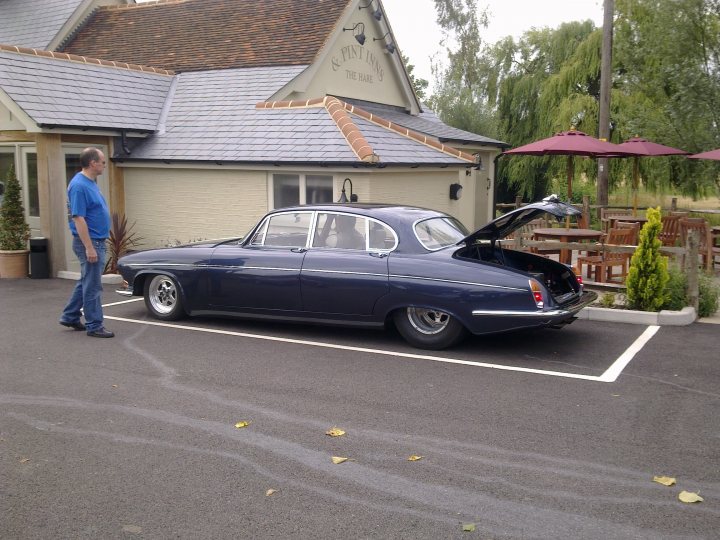 Pistonheads - The image captures a scene in a parking lot, with a man standing next to a blue vintage car. The car is parked at an angle, with its hood popped open, revealing the engine. The man, casually dressed in a blue shirt, jeans, and sneakers, seems to be inspecting the car. Behind them, a yellow building houses a restaurant or pub, recognizable by the chairs and tables visible through the windows. The environment appears calm, suggesting a leisurely day.