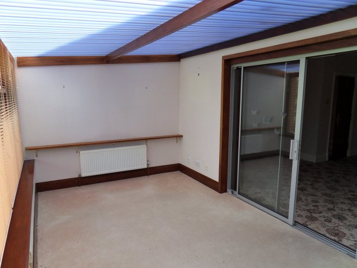 Replacing polycarbonate roof on converatory - Page 1 - Homes, Gardens and DIY - PistonHeads - The image shows an empty room with a wooden floor and a beige carpet meeting at a corner. Along one wall, there's a large window, which suggests plenty of natural light. The window is covered with white curtains, and on the opposite wall, there's a fireplace underneath a shelf, indicating a living area. The room has no furniture or decorations, which gives it a minimalistic and clean appearance.