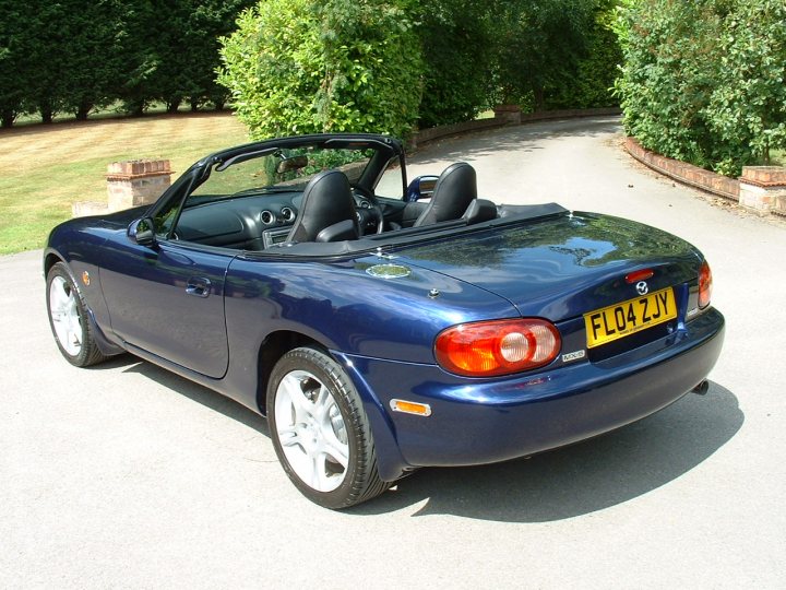 Pistonheads - The image shows a **blue convertible sports car** parked on a driveway. The car's top is down, suggesting it's a sunny day. The vehicle is situated on a gravel driveway, surrounded by lush greenery, indicating a residential area with mature landscaping. The front license plate reads "FLO4Y", and the car's fancy design adds to the impression that it's a stylish and well-maintained machine.