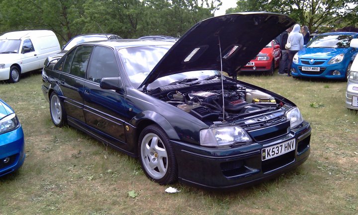 Cars On The Green - Photo Thread - Page 1 - East Anglia - PistonHeads - The image showcases a parking lot where several cars are neatly parked. The most prominent feature is a black two-door car with its hood propped open, revealing its engine. A group of people is gathered around this car, possibly members of a car club or mechanics discussing the vehicle. Surrounding this black car, other cars add to the diversity of the display, with varying colors and models. The setting seems to be an outdoor event or gathering, given the open sky visible in the background. There are no immediate texts or distinctive logos visible within the image.