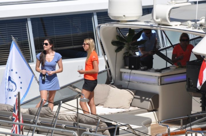 Celebrating 20 years of ... - Page 1 - Vipers - PistonHeads - The image captures a leisurely scene on a motorized boat. Two women, acting as hostesses, are standing on the deck, dressed in casual clothing. One is wearing a white dress and the other is in an orange shirt. They are holding beverage glasses suggesting they are serving drinks to the passengers aboard. The boat has an open top with a bar area, and the people on board appear to be enjoying a day out on the water.