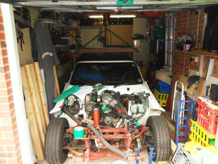 Body Lift - Page 1 - S Series - PistonHeads - The image shows an interior view of a garage, occupied by a white car positioned in the center of the frame. The car is being worked on or restored, as evidenced by the various engine parts and utensils scattered around the visible side. In the foreground, there is a red and white winch on a trailer, suggesting that the car may be undergoing a lifting or construction-related process in the garage. On the left, there's a grey garment hanging, and the walls of the garage are covered in various items, hinting at a work-intensive or hobby-focused environment.