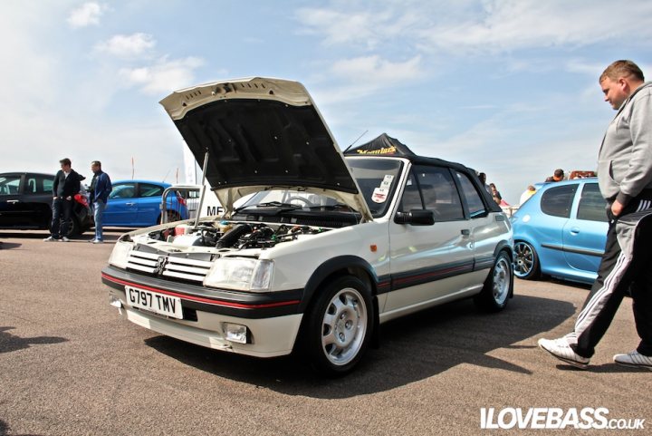 Anyone here know about 205 CTIs? - Page 1 - French Bred - PistonHeads - The image features an old white car with an open hood, parked in a lot filled with other vehicles. Numerous people are gathered around the car, one man appears to be examining the open hood of the white car while another man walks by, looking at the cars. The setting suggests an informal car show with individuals engaged in conversations and gleaning information about the vehicles.