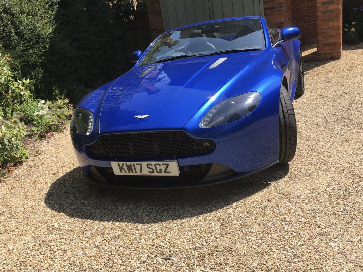 VANTAGE GRILLE .  - Page 2 - Aston Martin - PistonHeads - The image depicts a striking blue sports car parked on a driveway. The car, which is an Aston Martin Vantage, features a sleek and aerodynamic design with a prominent front grille. Its license plate reads "KW17 KGG". In the background, there's a hint of greenery and a house, suggesting a residential area. The image has a watermark or overlay that partially obscures the view of the car, making it slightly difficult to fully appreciate its details.