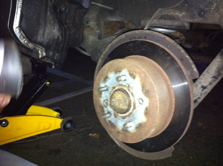 Hub Toyota Pistonheads Wheel Remove - The image shows a close-up of a brake rotor attached to a vehicle. The rotor is dirty and appears to have seen considerable use. It has numerous visible bolts that secure it to the brake caliper. A human hand is visible in the top left corner of the image, holding a socket tool, likely engaged in the task of changing the rotor or related maintenance. The surroundings suggest the setting is outdoors, likely on a road or a parking area.