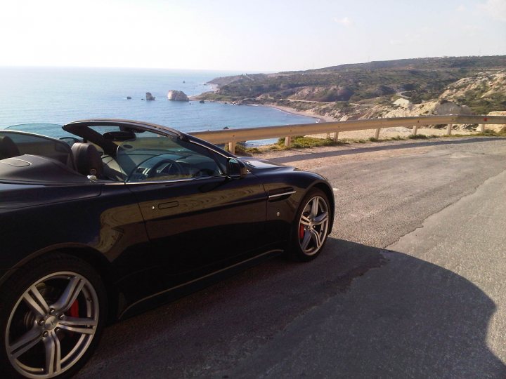 Hello from Cyprus! - Page 1 - Aston Martin - PistonHeads - The image showcases a striking black sports car parked on a road beside the ocean. The car is positioned close to the railing, possibly at a rest area or viewing spot. The ocean, visible through the car's windows, adds a serene backdrop to the scene. Behind the ocean, mountains can be seen in the background, hinting at a geographically diverse environment. The overall setting suggests a leisurely or holiday journey for the car owner.