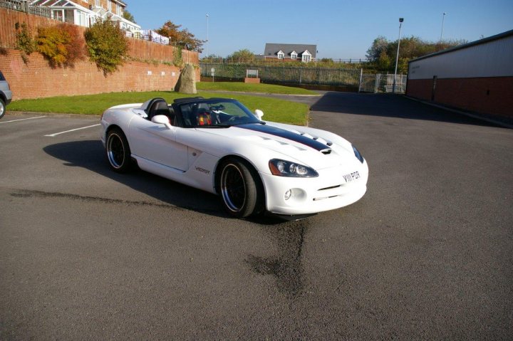 Rear Spoiler Or Not! - Page 1 - Vipers - PistonHeads - The image showcases a white sports car parked on an asphalt road next to a parking lot. The car, which appears to be a convertible, has its top down, revealing a sleek interior without the use of windows. In the background, there are residential structures, including houses and a tall brick building. A fence can also be seen dividing the parking lot from the residential area. The setting suggests a private or semi-private area, possibly a gated community or an area with restricted access.