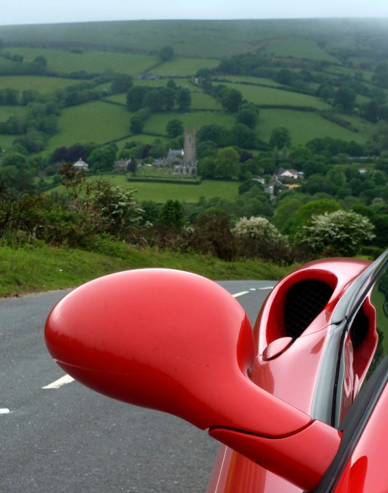 Isle of Arran in the Sunshine... - Page 1 - Supercar General - PistonHeads - The image presents a stunning view from the passenger side of a red car. The car's side mirror reflects a pastoral landscape dominated by vast fields and hills. The road beneath the car curves gently, with grass and shrubbery lining its sides. In the distance, farmhouses and a church are glimpsed, adding to the serene rural charm of the scene. The optimal angle of the shot provides a clear view of the road, the car, and the beautiful landscape in one frame.