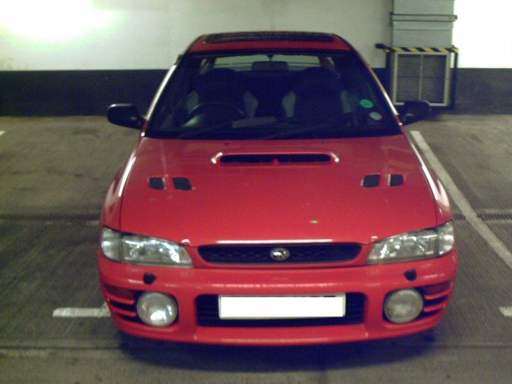 Impreza Pistonheads Turbo - This image shows a vibrant red sedan parked in a parking lot or parking garage. The car is facing towards the left side of the image, and it features two round headlights and a distinctive grille that suggest it's a model from the late 1990s to early 2000s. The license plate is clearly visible, it reads "**" and has an identifier with a number and a suffix. In the background, there is a wall and some lighting fixtures, indicating an indoor area.