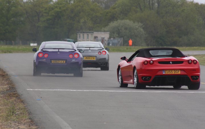 Pistonheads - The image depicts a scene on a road where a red sports car with yellow number plates, designated by "LEUBS DLF," is driving in the foreground. There are three cars in total: one grey car directly ahead of the red car, and a purple car following behind. The grey car has yellow license plates as well, and the purple car has a white license plate. The road appears to be an asphalt road, and the weather seems overcast.