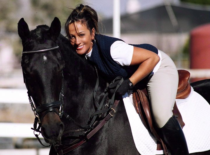 Importing my 2006 convertable mustang LHD from USA to NZ - Page 1 - Mustangs - PistonHeads - The image features a woman in a blue shirt, plaid vest, and a ponytail, smiling while horseback riding. She is wearing riding clothes, including black gloves. Behind her, a green, grassy field is visible, giving the impression of a countryside riding scene. She is seated upright on a black horse with a shining coat, harness, and bridle, indicating that it is well-cared-for and possibly serves as a fixture in equestrian events or trail riding.