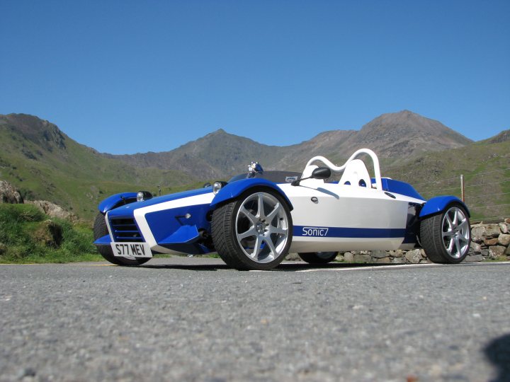 Pictures of your Kit Car..? - Page 22 - Kit Cars - PistonHeads - The image features a small, blue race car with white accents, parked on an asphalt surface. The car is positioned to show a clear view of its front wheels. Rolling foothills rise in the background, suggesting a highland environment, which complements the overall interesting design of the car. The car appears to be a one-seater, likely designed for speed and agility. There is an open sky above, which makes the scenery even more picturesque.