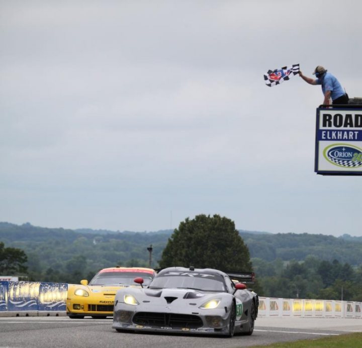 Pistonheads - The image captures a thrilling moment at the RR spinning road competition at Road America. In the foreground, a silver race car zooms past, embodying the speed and edge of the action sports scene. Further back, another yellow car is visible, perhaps a competitor following the leader. 

Above the scene, a flagger stands poised, waving his checkered flag, a universal symbol of conclusion in many sports including racing. The high angle of the shot gives a dramatic view over the race, providing a sense of the bustle and adrenaline of the race track. The background is a lush green, possibly the grass or stage area of the track, contrasting with the fast-moving cars.