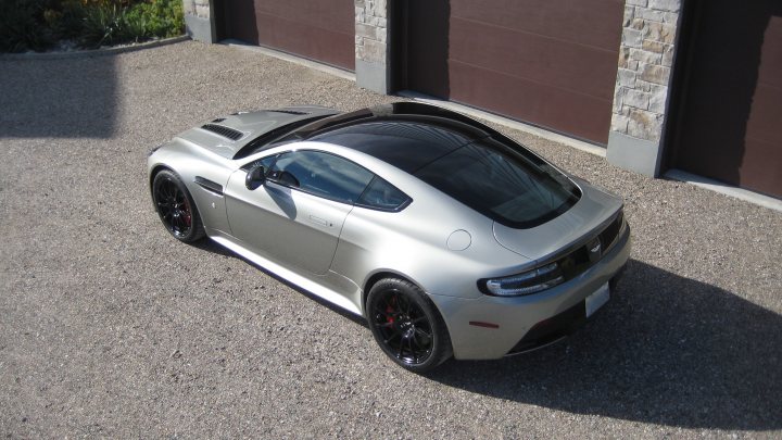 Best wheels for silver cars [submit pics] - Page 1 - Aston Martin - PistonHeads - The image showcases a silver sports car parked on a gravel driveway adjacent to a tall red brick fence. The car presents a sleek, low-profile design with a distinct angular rear profile. Its dynamic styling stands out against the warm brickwork and the neutral tones of the gravel surface. The car is partially in the shadow, adding a sense of depth and interest to the composition. The rocky landscape in the background provides a natural contrast to the man-made objects and the urban feel hinted by the red brick fence.