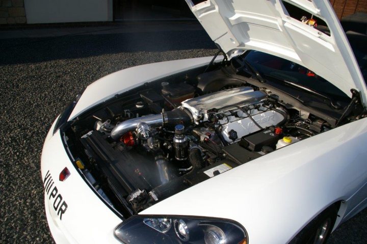 Blown But Not Blown - Page 1 - Vipers - PistonHeads - The image shows a white car with its hood open, revealing a complex mechanical engine underneath. The engine has various metal components, including pipes, belts, and moving parts typical of a car engine. Above the engine, a silver or chrome metal cooler tank is prominently visible, reflecting the light in the photo. A red light on the car's back is also reflected in the photo. The car is parked on an asphalt surface, indicating a possible maintenance or inspection activity might be taking place.