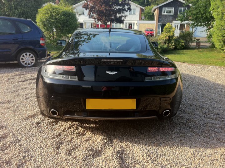 Changed Spot Pistonheads - The image shows a dark-colored sports car parked on a gravel driveway in front of a residential house with a white exterior and a garage. The car appears to be a new model with a sleek design, featuring a license plate at the rear. The setting includes a well-maintained lawn and a tree, suggesting a suburban or rural environment. The vehicle has a distinctive taillights with red lights and two round shapes above the back lights. There is reflective white on the door handles and the side view mirrors, which is a feature commonly found on luxury or sports cars.