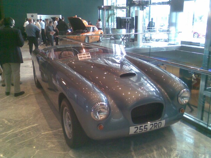 Wharf Canary Motorexpo Interesting Pistonheads - The image showcases an open car show in a well-lit room with reflective marble floors. A silver-colored convertible sports car, likely a classic or vintage model, is displayed prominently. It's parked perpendicular to the viewer's perspective, with its large hood open, revealing the engine. The vehicle has a license plate, and there are brochures and signage near it, including a placard on the windshield that seems to provide information about the car. The audience, composed of individuals in various attires, surrounds the car, observing it intently. The overall setting suggests a dedicated flagship motor racing store or a lavish private gallery where the car is displayed for viewing or purchase.