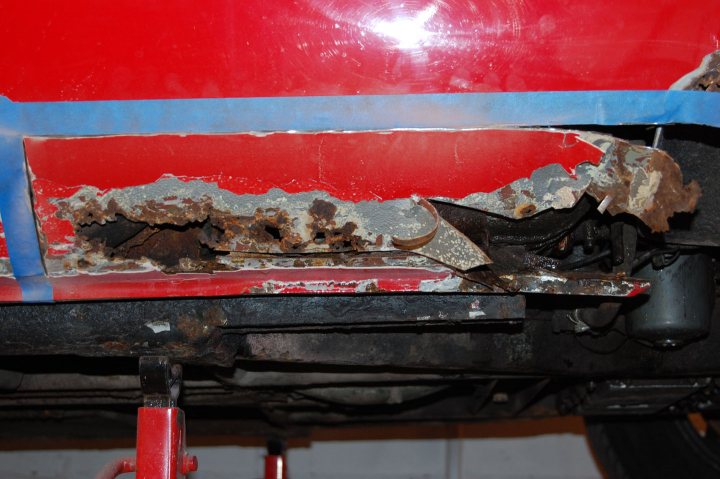 Classic Supercar Pistonheads Experiences - The image shows a close-up view of a piece of machinery, specifically the back of a red vehicle. The machine component appears to have heavy usage, with rust becoming a problem area. Visible damage includes rust, chipping, and what seems to be a crack or structural issue along the side and back surfaces. A blue tag with a white stripe is attached to the component, indicating caution or attention to repair issues.