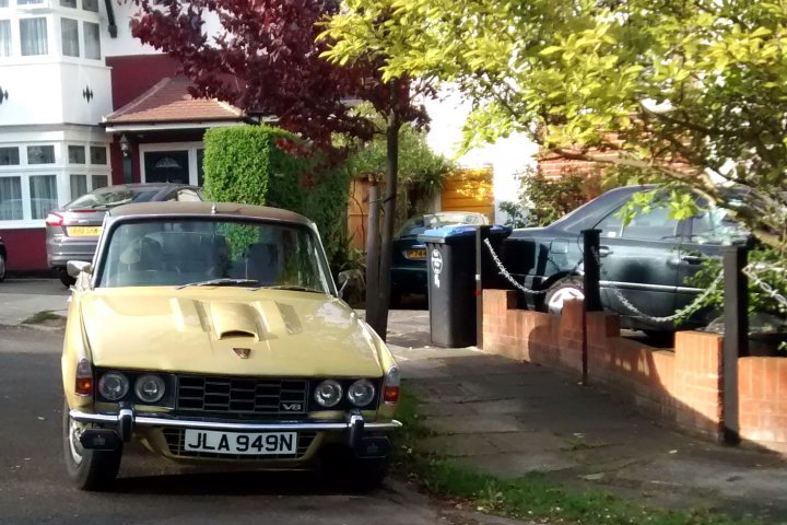 A car is parked on the side of the road - Pistonheads - The image depicts a narrow urban street scene. A yellow and black vintage car is parked on the left side of the street. The car is old-fashioned with round headlights and a trunk cover. The backdrop features two trees that partially obstruct the view of the building behind. A wooden fence runs along the right side of the street, providing some separation from the adjacent properties. The surrounding environment suggests a residential area with parked cars and greenery.