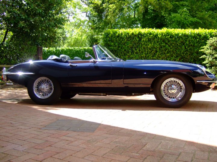 Get your Jags out - Show us your car - Page 4 - Jaguar - PistonHeads - The image shows a classic car parked on a brick pavement under a clear sky. The car is of old luxury design with a convertible top, evident from the absence of a roof. It features wire wheels and a gleaming exterior, reflecting the sunlight. The background is a lush, well-maintained garden with verdant shrubbery and mature trees contributing to a serene and idyllic setting. The overall style of the image is bright and sunny, capturing the car in a pleasant contrast to the natural elements.