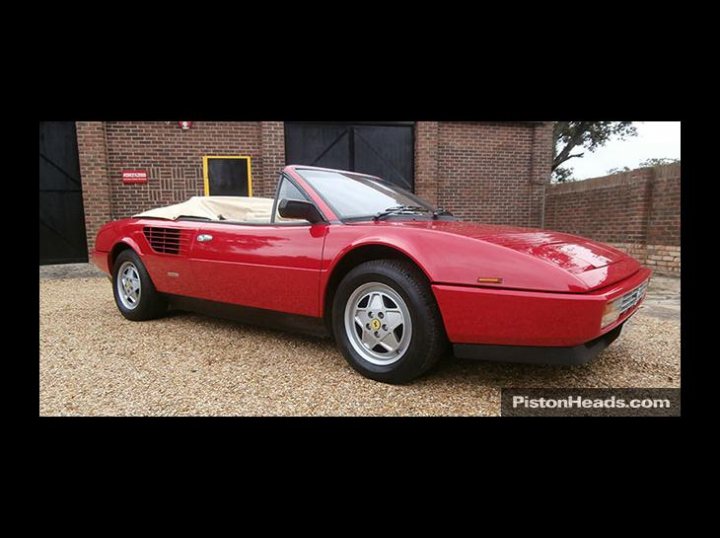 Don't you just love PH - Page 1 - Australia - PistonHeads - The image features a red Ferrari sports car parked on a pebble-covered ground. The car is iconic with its sleek, open-top design and distinctive black and silver rims. The seats are visible and appear to be red as well, matching the overall color scheme of the vehicle. The surroundings suggest an indoor setting, possibly a showroom or exhibition space, with a brick wall visible in the background.