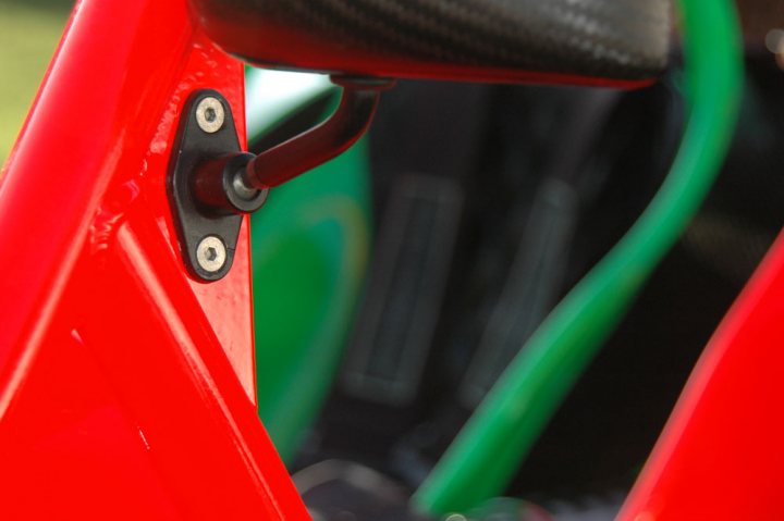 Spa Mirrors Pistonheads Fitting Rollcage - This is a close-up photograph focusing on the red-toned body of a vehicle, taken at an angle where a part of a metallic bar or structure is visible despite being mostly obstructed by a black object, which could be a standard clamp, typical of machinery or vehicle components. The black object is attached to the red structure, partially leaning to one side. Beyond the structure, the background is bright and blurred, suggesting it might be outdoors. The phrase "the other side of the world" is also visible in the image, possibly indicating a new setting, trip, or concept related to global exploration or cultural exchange. The style of the image is realistic, and the quality of the photograph suggests it might have been taken to showcase the details of the vehicle or the part of the tool that is clamped.