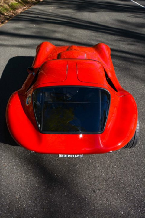 Bill Thomas Cheetah - Page 1 - General Gassing - PistonHeads - The image shows a close-up of a sleek red sports car, designed with a streamlined body, potentially inspired by the concept of aerodynamics for increased speed. The car is parked on an asphalt street, with a portion of its numberplate visible, suggesting a license plate number that is not entirely clear. The car's body reflects the hues of the environment, particularly a hint of green, possibly indicating the surrounding vegetation, and the grids are consistent, implying a well-maintained surface. The overall composition of the image highlights the agility and presence of the sports car on a quiet, clean street.