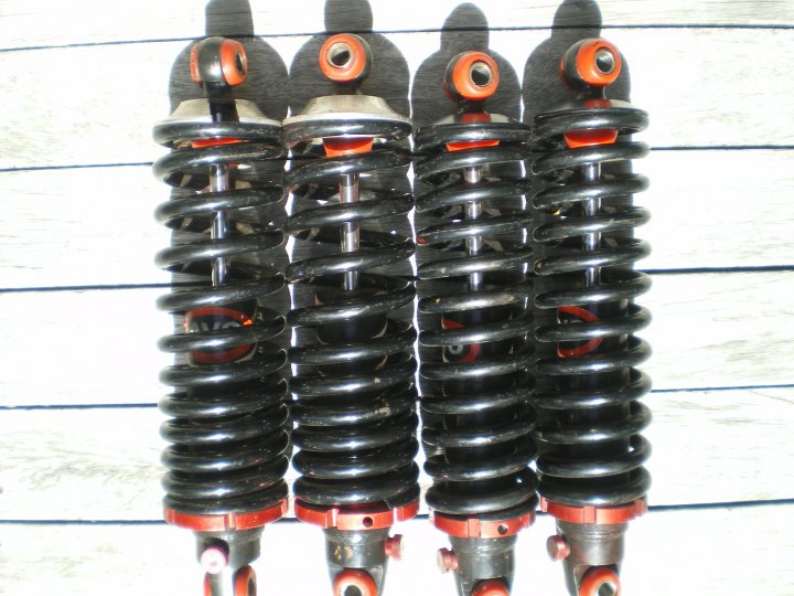 Avosprings Question Pistonheads - The image displays a set of three automobile suspension coils against a wooden plank background. These coils appear to be from a car, likely removed from a vehicle for maintenance. They are black with red accents at their ends and screwed onto bearings at both ends. The background shows horizontal stripes of wood and a uniform white surface, likely painted wall or board. The lighting focuses on the coils themselves, highlighting their texture and material.