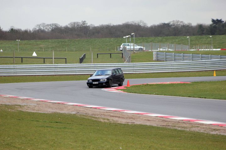 Track day at Snetterton Friday 15th February - Page 1 - East Anglia - PistonHeads - The image shows a gray, four-door car navigating a left turn on a race track. The car is on its right side, suggesting it is in a non-English driving country. Behind the car is a beige safety car, possibly its escort, and there is a red cone indicating a temporary boundary or hazard on the track. The race track is surrounded by grass, indicating that it's an outdoor circuit, and there are a few spectators in the background, looking towards the safety car. The sky is overcast, and the overall scene conveys a sense of motion and testing.