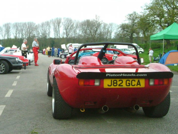 Seized Engine Pistonheads - In the center of the image, a vibrant red sports car takes the stage on a parking lot with white markings. The license plate of the car reads "J82 GCA," indicating a connection to a masquerade event. The shiny body of the car reflects the surroundings, and a yellow license plate stands out against the red. There are numerous people in the background, some standing and others near various vehicles, contributing to the lively atmosphere of a gathering, possibly a sports car gathering or a masquerade event. The scene suggests a gathering of car enthusiasts in the daytime.