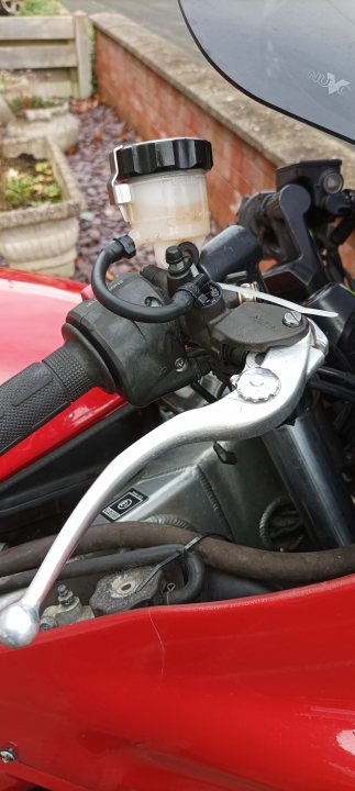 Pistonheads - This image shows a red motorcycle parked, with its engine exposed. The motorcycle is set against a backdrop that includes what appears to be a brick wall and some greenery. On the right side of the photo, there's an overlaid graphic resembling a person's face, creating an illusion as if a face were emerging from the engine compartment. This gives the impression that the motorcycle is "talking" or has a human-like presence within it. The overall image conveys a creative and whimsical tone.