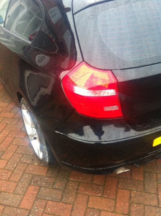 Will this be a write off? Estimated repair cost? - Page 2 - BMW General - PistonHeads - The image captures a close-up view of a black car parked on a brick driveway on a rainy day. The focus of the image is on the rear left tire of the car and its reflection on the polished surface of a nearby mirror. The wet city street subtly mirrors the car in the distance. The car features a unique design with a red dot on the side, adding a pop of color to the otherwise monochromatic scene. The rain leaves a slight sheen on the car, contributing to the overall mood of the image.