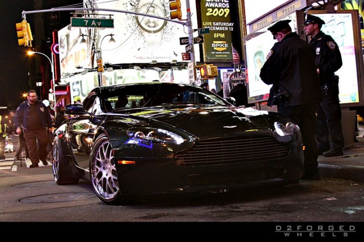 Car Porn - Page 1 - Aston Martin - PistonHeads - The image depicts a nighttime street scene, featuring a Bentley car parked by the side of the road. Two police officers in uniform are standing nearby, engaged in conversation with a civilian. The street is illuminated with neon lights and billboards adorn the background. The image has a credit at the bottom indicating it may have been created or edited by or associated with 'D2FORGED WHEELS.'