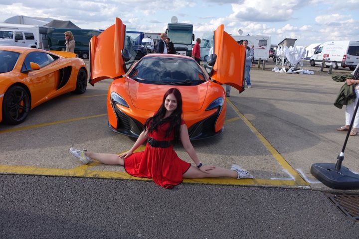 Silverstone Classic 2016 - McLaren Drivers Club - Page 1 - McLaren - PistonHeads - The image captures a dynamic scene in a parking lot where a stunning woman is striking a pose in front of a distinctively colored car. The car, painted in a vibrant shade of orange, is equipped with a gargantuan wing that sets it apart from others. The parking lot is bustling with activity, filled with various vehicles, and there are a multitude of people attending to their cars. The woman in the red dress exudes confidence, adding a splash of color to the scene. The image is full of movement and energy, encapsulating the lively atmosphere of the parking lot.