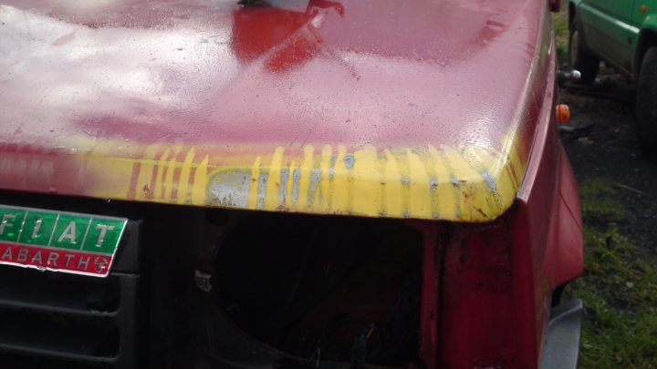 An old suitcase is sitting on the ground - Pistonheads - The image shows the rear of a vehicle, likely a truck, with a prominent spare tire carrier. The carrier's body appears to be fading yellow with a rust-like texture, indicating it has seen significant use or exposure to the elements. There is a red stamp painted on it, and underneath, the label "FIAT" and "ABARTHS" are visible, suggesting the model or brand of the vehicle. The overall backdrop is out of focus with greenery, possibly plants or grass, indicating the photo might have been taken outdoors, possibly in a garden or yard.