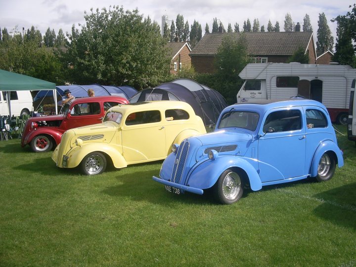 Steel Pistonheads Yank Cruisers City - The image captures a lively scene of a vintage car show in a park. There are four classic car models on display, each boasting a unique color and design. One is a vibrant red, another a cheerful yellow, a third a deep blue, and the last a classic white. These cars are stationed on the lush green grass, adding to their vintage charm. The backdrop is filled with a suburban landscape, creating a picturesque setting for this automobile gathering. Among the attendees, one individual is seen approaching the yellow car, possibly intrigued by its classic design.